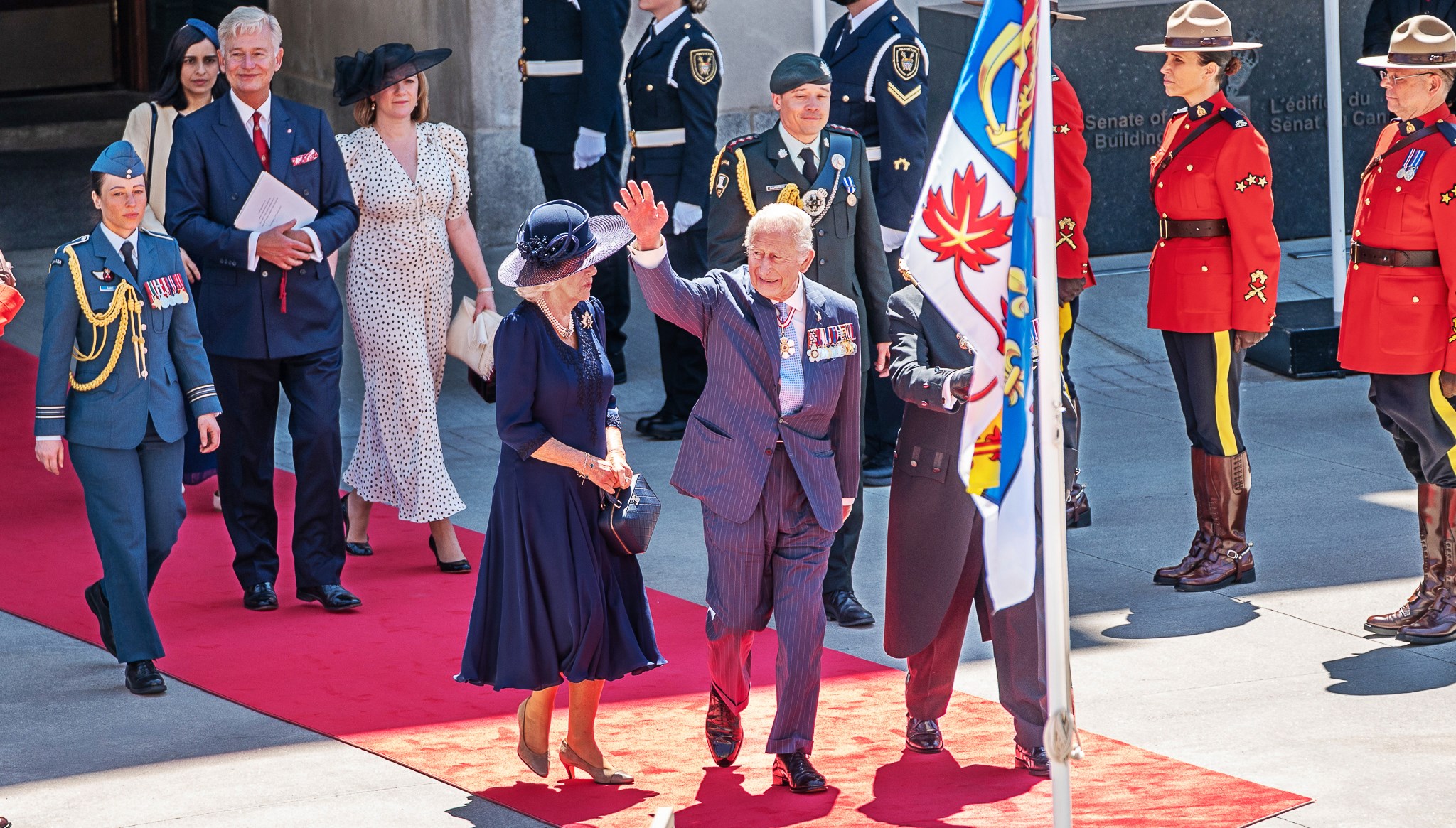 Le roi Charles III salue la foule de la main à partir du tapis rouge, devant l’édifice du Sénat du Canada; on peut voir le drapeau canadien personnel du monarque au premier plan.